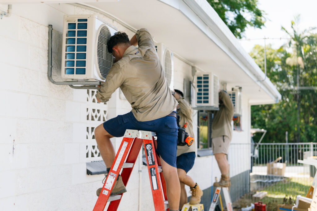 Technician on a step ladder servicing outdoor condenser units mounted along a wall