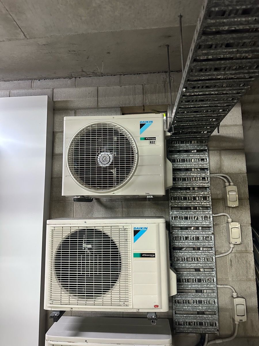 Two outdoor air conditioning condenser units stacked on wall brackets in a commercial plant space